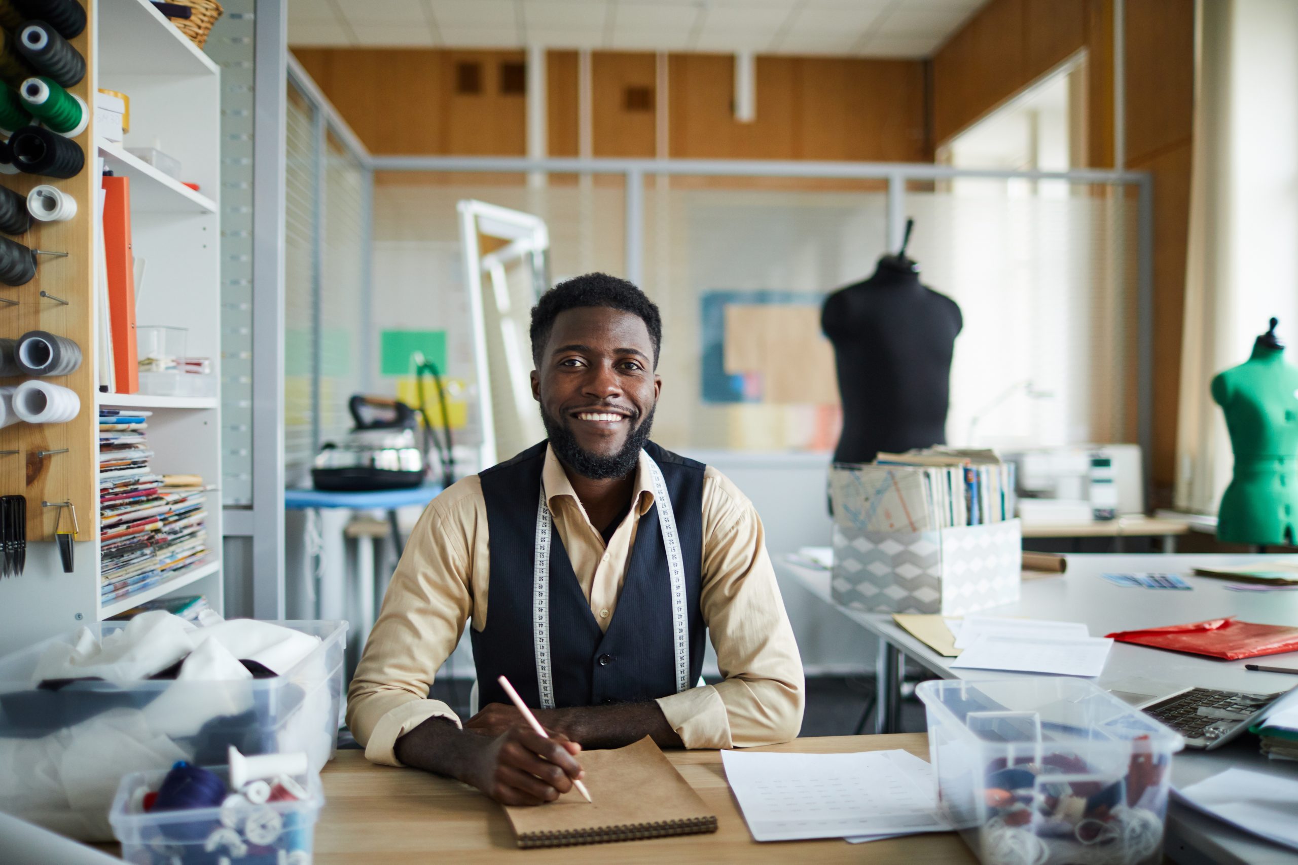 Young cheerful fashion designer sitting by table in his workshop and making new sketches in notepad